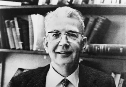Black and white portrait of a smiling man with glasses in front of bookshelves.