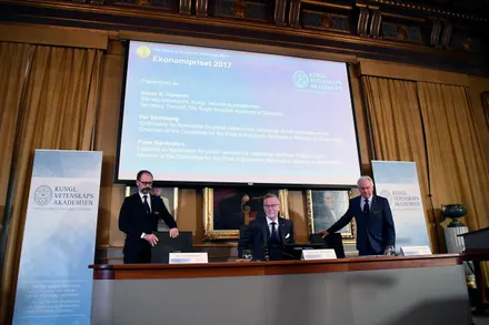 Three men in suits stand and sit before a large presentation screen in a formal room.