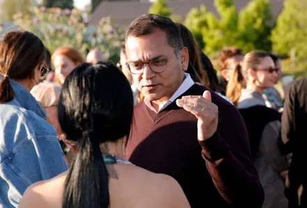 Man in brown sweater gesturing while talking to a woman outdoors.
