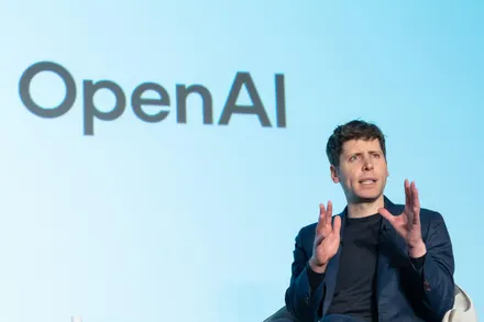 Person speaking at OpenAI event, gesturing hands.
